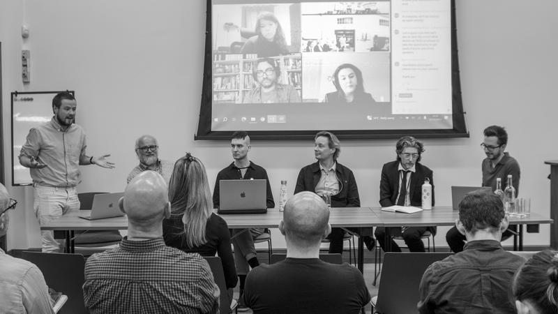 Black and white photo of the panel sat at a table in front of a projector screen showing remote attendees. 
