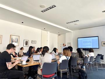 A group of summer school students sit around a table using laptops, a screen in the background shows a slide