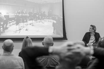 Joris de Man stands at a lectern in front of a projector screen showing musicians playing a piece