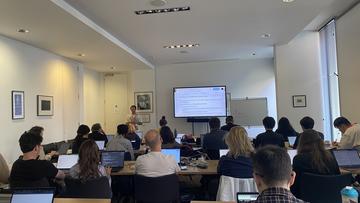 A photo taken from the back of a classroom - several people face towards a TV showing a slide titled 'Selecting and Inspecting Tabular Data', the instructor stands next to the screen