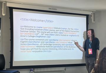 Huw stands in front of a projector pointing to a slide that is written in marked up text